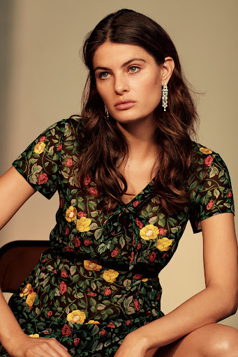 Portrait of a woman wearing a black floral print sheer dress and ornate pendant earrings against a warm neutral background.