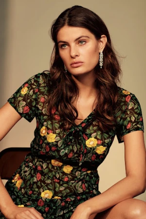 Portrait of a woman wearing a black floral print sheer dress and ornate pendant earrings against a warm neutral background.