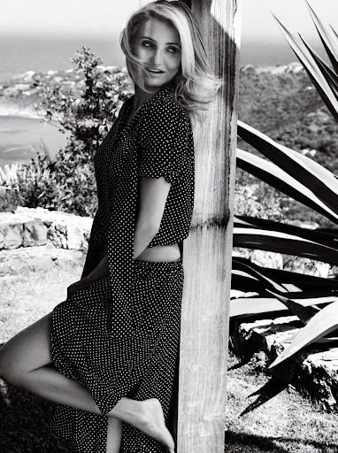 Black and white portrait of an adult woman with long blonde hair in a polka dot dress leaning on a wooden structure outdoors.