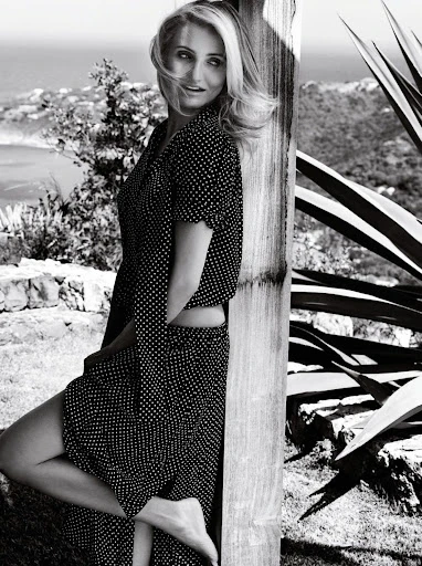 Black and white portrait of an adult woman with long blonde hair in a polka dot dress leaning on a wooden structure outdoors.