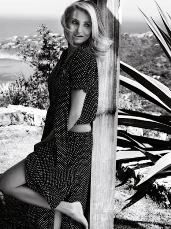 Black and white portrait of an adult woman with long blonde hair in a polka dot dress leaning on a wooden structure outdoors.