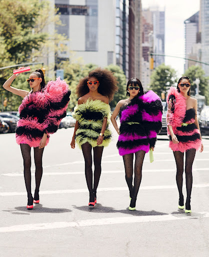 Four fashion models stride across a city crosswalk in bright, feathered mini-dresses with matching heels and patterned tights.