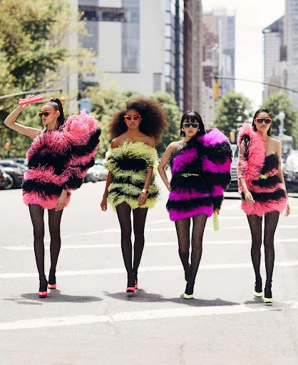 Four fashion models stride across a city crosswalk in bright, feathered mini-dresses with matching heels and patterned tights.