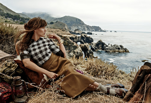 Woman in a checkered top and brown skirt sits on a hillside overlooking the Pacific Ocean, with camping gear and coastal mountains.