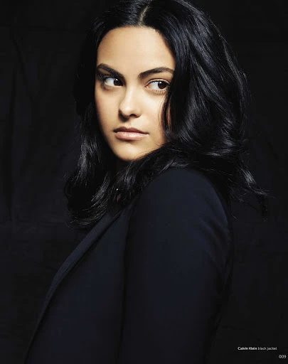 Portrait of adult woman with dark hair and navy blazer looking sideways against a dark background in a studio setting.