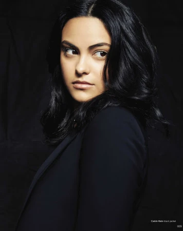 Portrait of adult woman with dark hair and navy blazer looking sideways against a dark background in a studio setting.