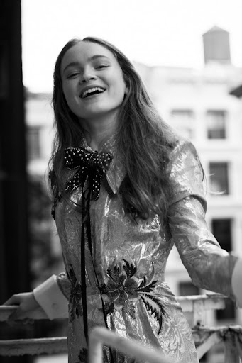 Portrait of a woman in a metallic floral dress with a large bow, smiling against a building backdrop in a black and white editorial photograph.