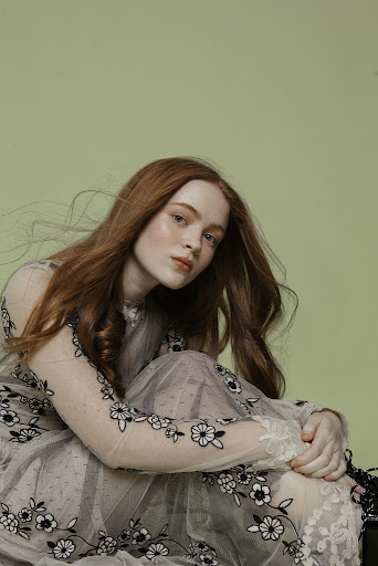 Portrait of a red-haired woman in a floral-embroidered dress, seated in a photo studio with a light green background, looking at the camera.
