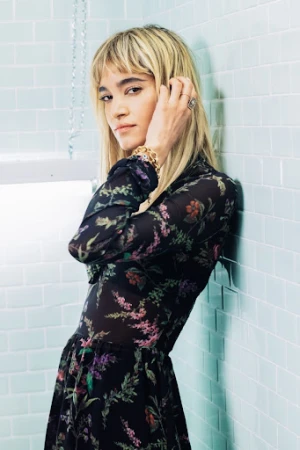 Young woman with blond hair leans against tiled wall in floral print dress, accessorized with jewelry, in a fashion portrait.