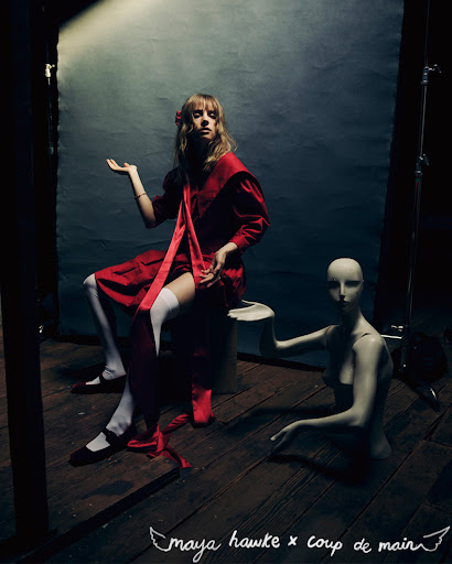A young woman in a red dress sits on a stool next to a mannequin in a dimly lit space with a distressed backdrop and wooden floor.
