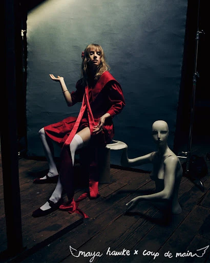 A young woman in a red dress sits on a stool next to a mannequin in a dimly lit space with a distressed backdrop and wooden floor.