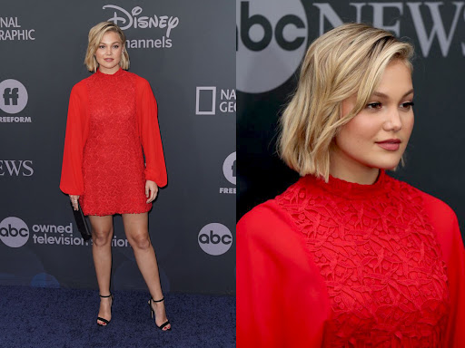 Olivia Holt attends an event, wearing a red lace dress and black high heels, smiling against a branded backdrop. Celebrity fashion and style.