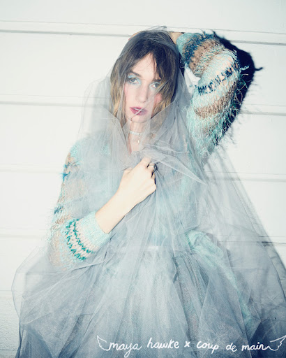 A person in a patterned sweater stands against a white wall with a tulle veil, posing for a fashion editorial with soft, diffused lighting.