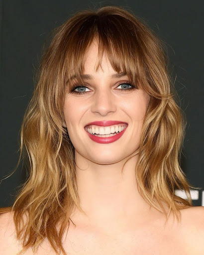 Close-up portrait of a smiling young woman with blonde wavy hair and bangs, wearing red lipstick against a dark background.