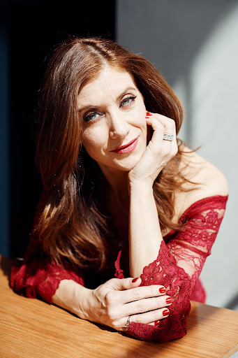 Close-up portrait of a mature woman with auburn hair, wearing a red lace top, resting her head on her hand, with warm lighting.