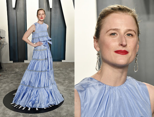 Woman in layered, light blue evening gown at a formal event, accessorized with drop earrings and red lipstick, standing against a neutral backdrop.