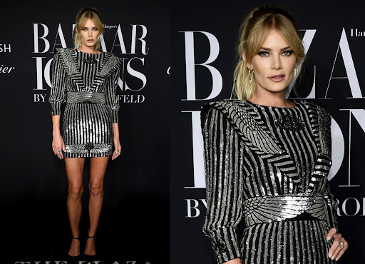 An adult woman poses on a red carpet at an event wearing a black and silver sparkling dress. She is standing in front of a branded backdrop.