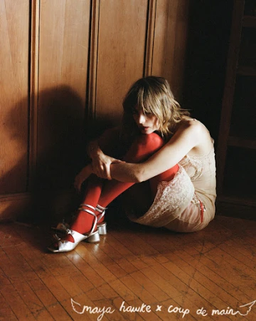A young woman in vintage style sitting against a wood panel wall wearing a lace dress, red tights and silver shoes with a distressed look.