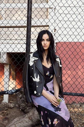 Portrait of a brunette woman with a black leather jacket draped over her shoulders, sitting outdoors against a chain-link fence.