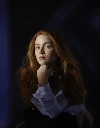 Portrait of young woman with long red hair wearing a light blue collared shirt and dark vest, with her chin resting on her hand.