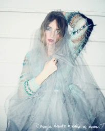 Maya Hawke wearing a light blue textured sweater and grey tulle veil against a white wood panel background.