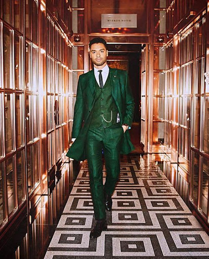 Stylish man in green suit walks down hallway with mirrored walls and patterned floor, posing for a fashion shoot in an indoor setting.
