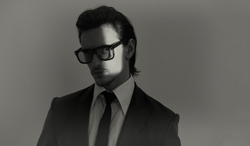 Portrait of a man wearing dark glasses, a suit, and tie against a plain background with dramatic lighting across his face.