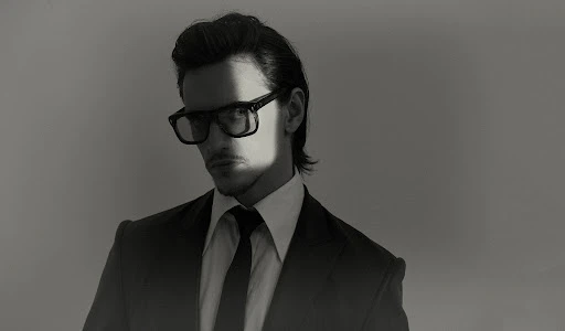 Portrait of a man wearing dark glasses, a suit, and tie against a plain background with dramatic lighting across his face.