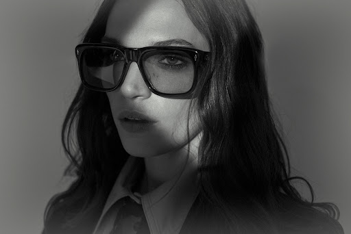 Close-up portrait of a woman wearing black square sunglasses and dark collared shirt against a grey background
