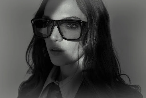 Close-up portrait of a woman wearing black square sunglasses and dark collared shirt against a grey background