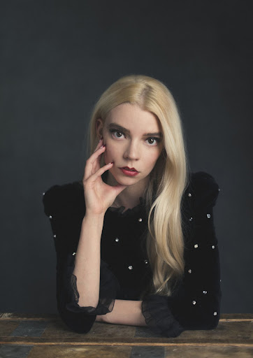Close-up portrait of a blonde woman with red lipstick wearing a black dress with pearl embellishments sitting in front of a gray background.