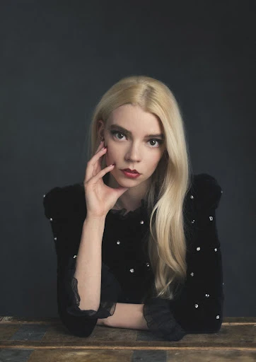 Close-up portrait of a blonde woman with red lipstick wearing a black dress with pearl embellishments sitting in front of a gray background.