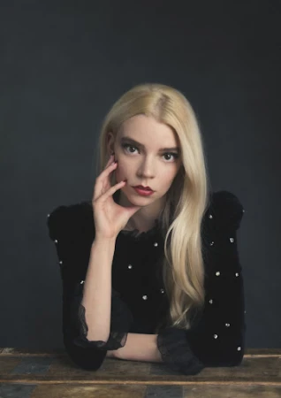 Close-up portrait of a blonde woman with red lipstick wearing a black dress with pearl embellishments sitting in front of a gray background.