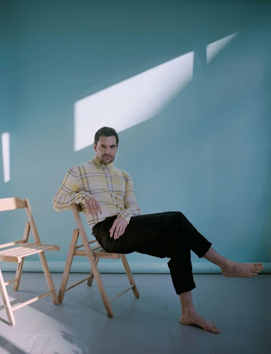 Portrait of a man in a yellow plaid shirt and black pants sitting cross-legged on a wooden chair against a blue wall with sunlight.