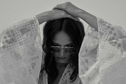 Monochrome portrait of a woman wearing sunglasses and a patterned white cover-up, looking down with her hands above her head.