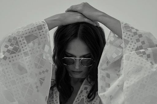 Monochrome portrait of a woman wearing sunglasses and a patterned white cover-up, looking down with her hands above her head.