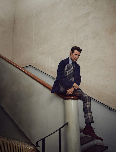 A stylish man in a blue blazer and plaid pants sits on a staircase, indoors, in a fashion-forward pose with soft, diffused lighting.