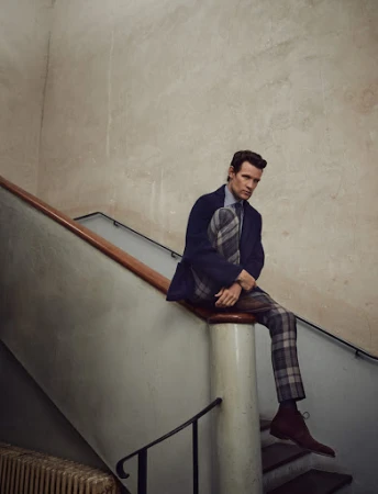 A stylish man in a blue blazer and plaid pants sits on a staircase, indoors, in a fashion-forward pose with soft, diffused lighting.