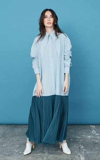 Full-length portrait of a woman wearing a light blue oversized shirt and dark green pleated maxi skirt against a solid blue backdrop.