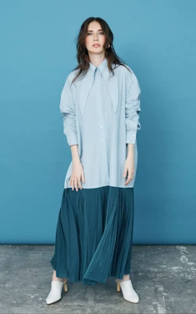 Full-length portrait of a woman wearing a light blue oversized shirt and dark green pleated maxi skirt against a solid blue backdrop.