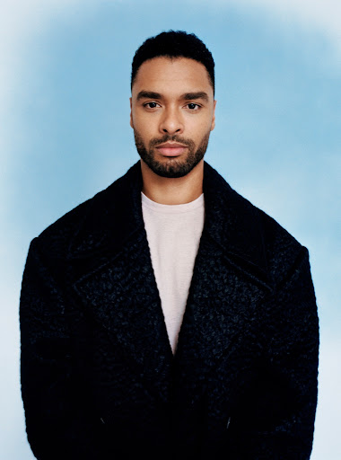 Regé-Jean Page portrait in a dark coat against a bright sky blue background, looking directly at the camera with a neutral expression.