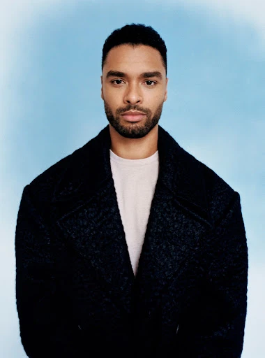 Regé-Jean Page portrait in a dark coat against a bright sky blue background, looking directly at the camera with a neutral expression.