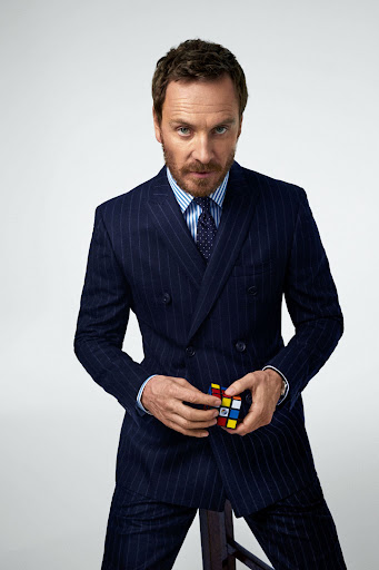 A stylish adult male in a pinstripe suit is holding a Rubik's Cube in a portrait shot against a neutral studio backdrop with soft, even lighting.