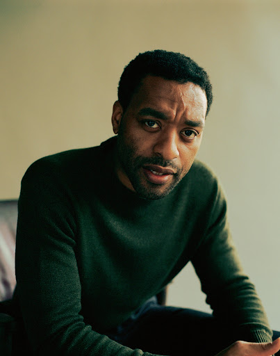 Portrait of actor Chiwetel Ejiofor in an olive green sweater, seated against a neutral backdrop in a studio setting with natural lighting.