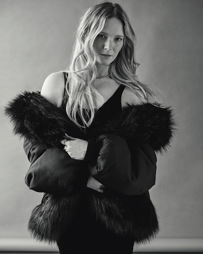 Monochrome portrait of a blonde woman in a black top and black coat with fur trim against a plain gray background, looking at the camera.