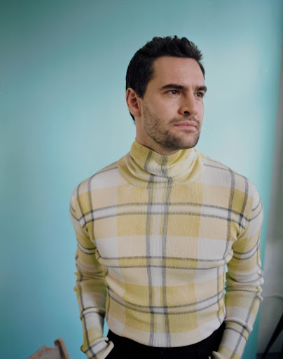 Man wearing a yellow and white plaid turtleneck sweater against a light blue backdrop.