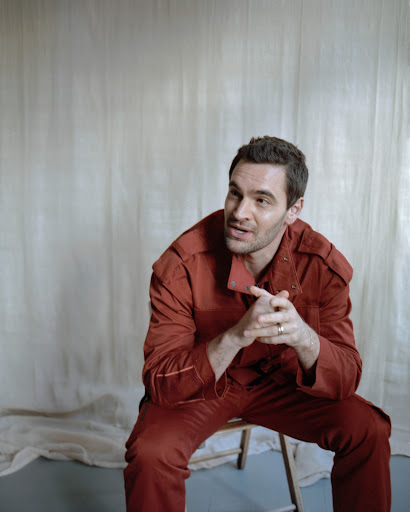 Portrait of a dark-haired adult man in a red jumpsuit sitting on a wooden chair against a neutral backdrop in a photo studio.