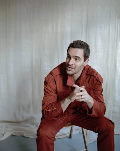 Portrait of a dark-haired adult man in a red jumpsuit sitting on a wooden chair against a neutral backdrop in a photo studio.