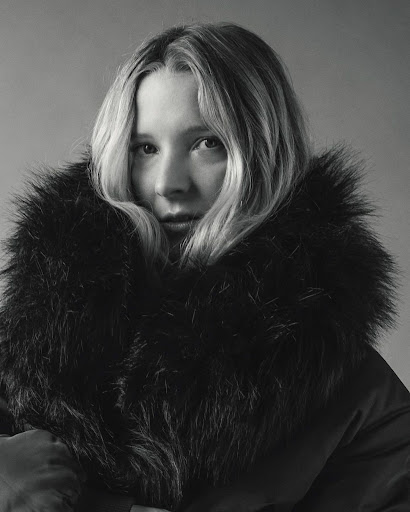Portrait of a blonde woman wearing a fur-trimmed winter coat against a neutral backdrop, emphasizing fashion and studio photography.