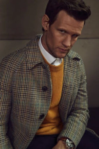 Portrait of a man in a plaid overcoat and mustard-colored sweater. He is posed indoors, looking toward the camera with a slight smile.
